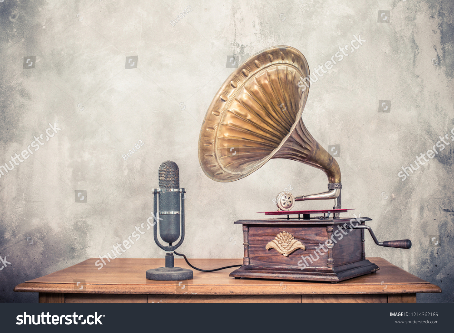 Vintage antique gramophone phonograph turntable with brass horn and big aged studio microphone on wooden table front concrete wall background. Retro old style filtered photo #1214362189