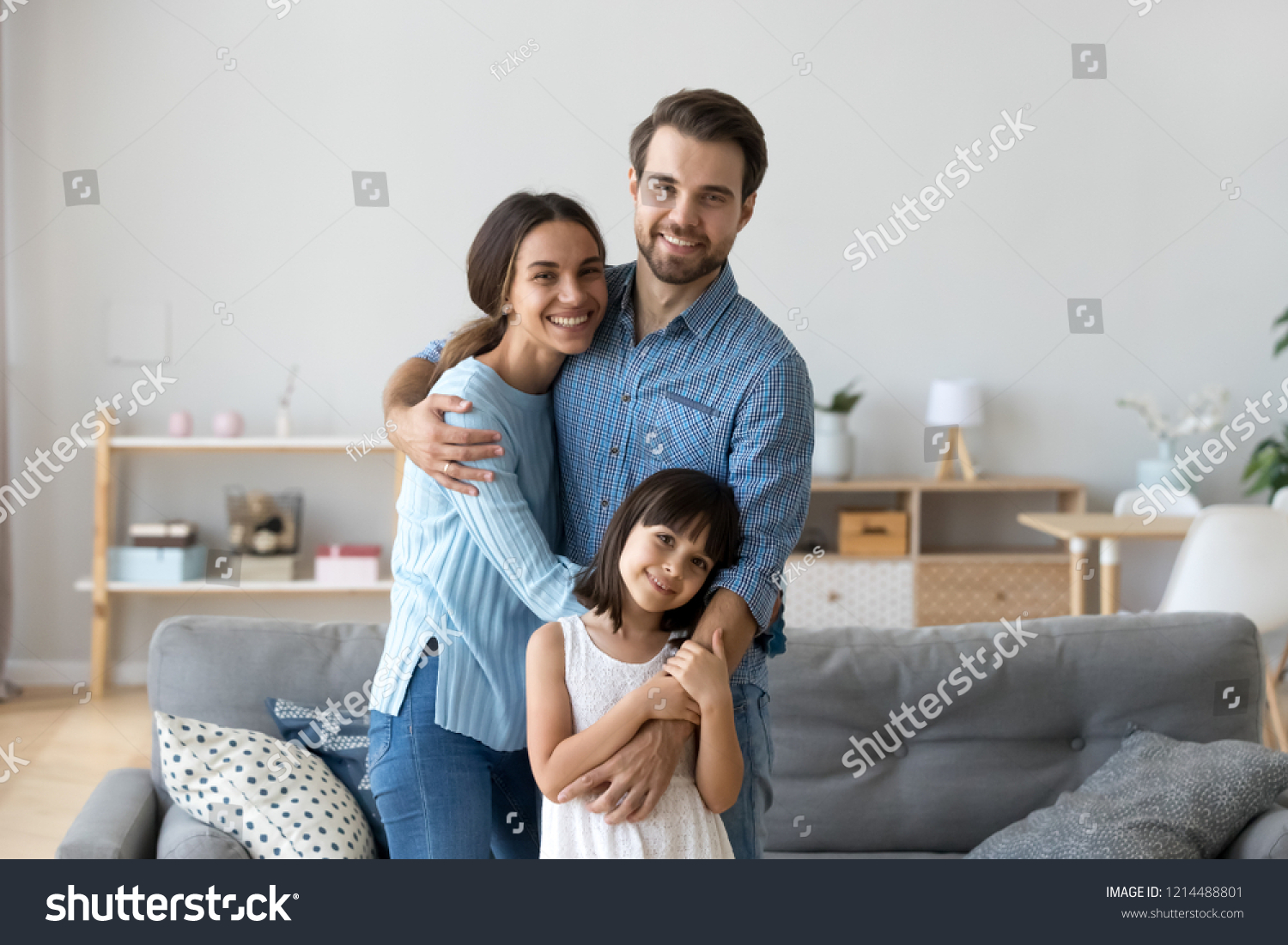 Cheerful diverse multi-ethnic family married couple wife husband little daughter embracing standing together in living room smiling looking at camera at new modern home feels happy and satisfied #1214488801