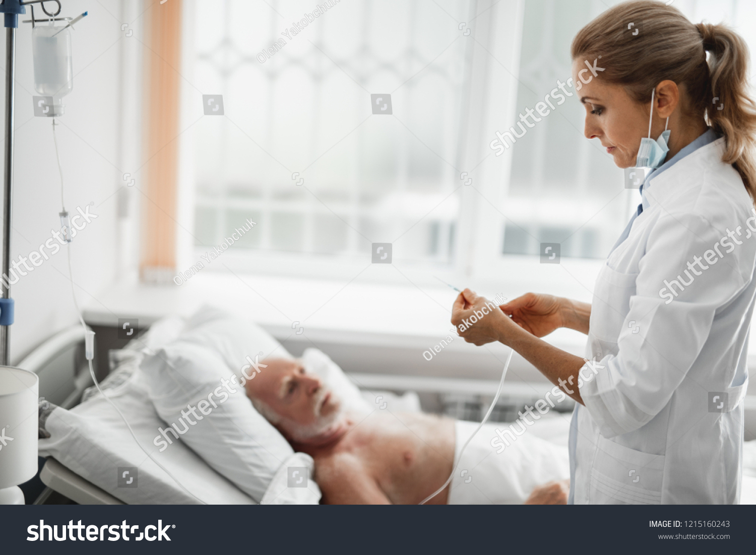 Side view portrait of young lady in white lab coat holding needle for intravenous infusion. Old man lying in bed on blurred background #1215160243