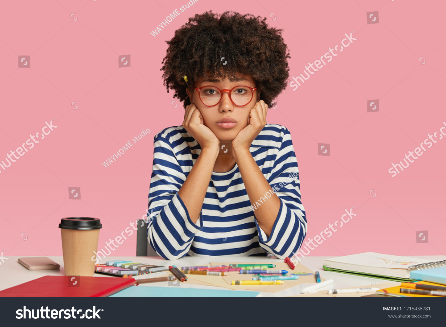 Sad dejected female architect feels unhappy as has no inspiration for creating masterpiece, sits at desktop, surrounded with crayons, notebook and disposable cup of coffee, holds chin isolated on pink #1215438781