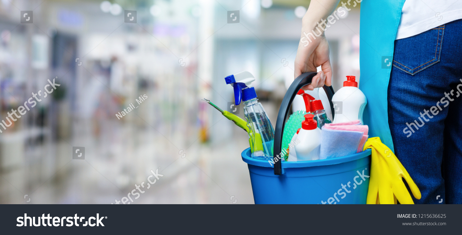 A cleaning woman is standing inside a building holding a blue bucket fulfilled with chemicals and facilities for tidying up in her hand. #1215636625
