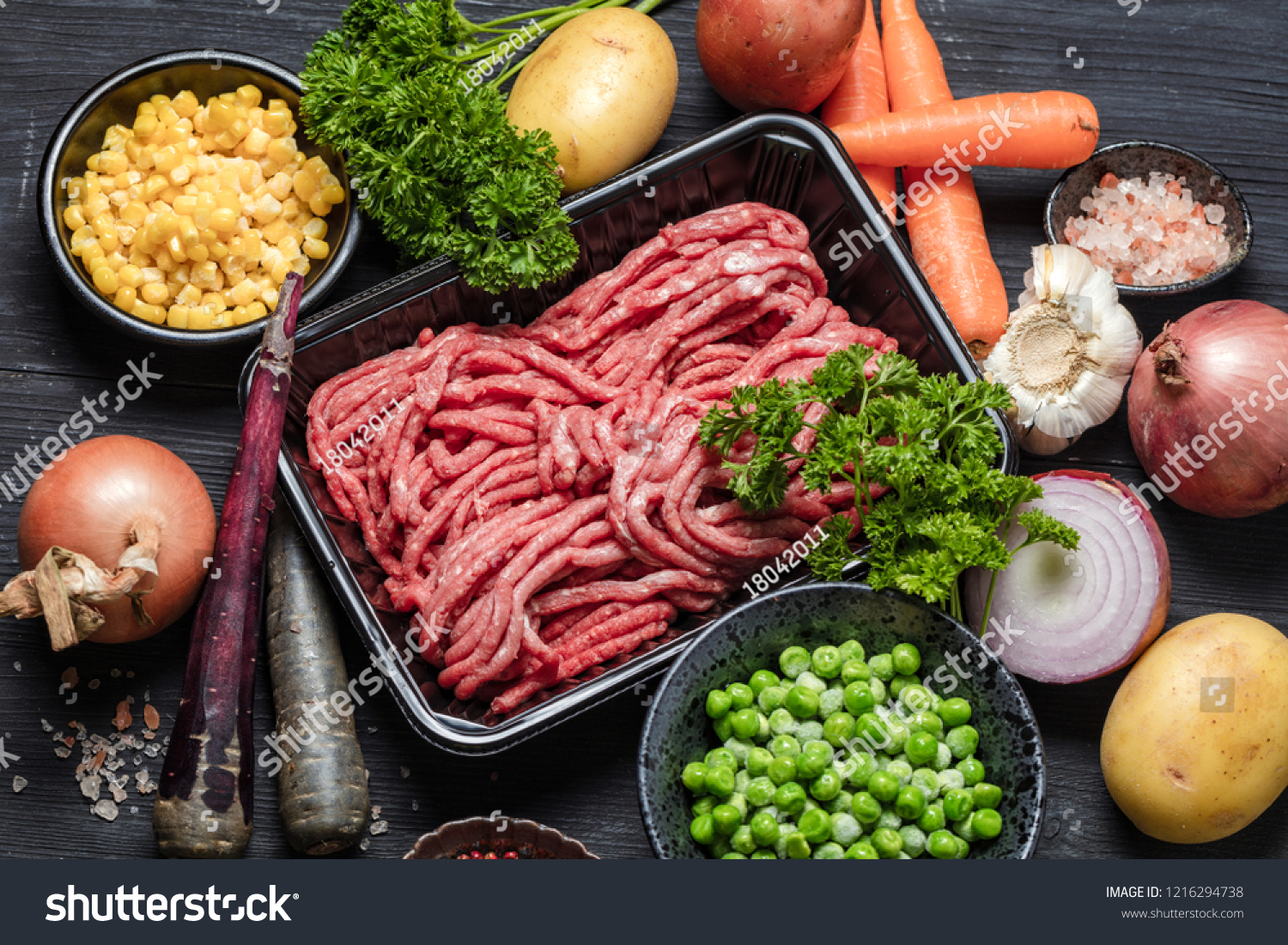 Raw minced meat in container surrounded by ingredients for shepherds pie , green peas, yellow corn, carrot, onion and seasonings, on black background, top view. Horizontal composition  #1216294738