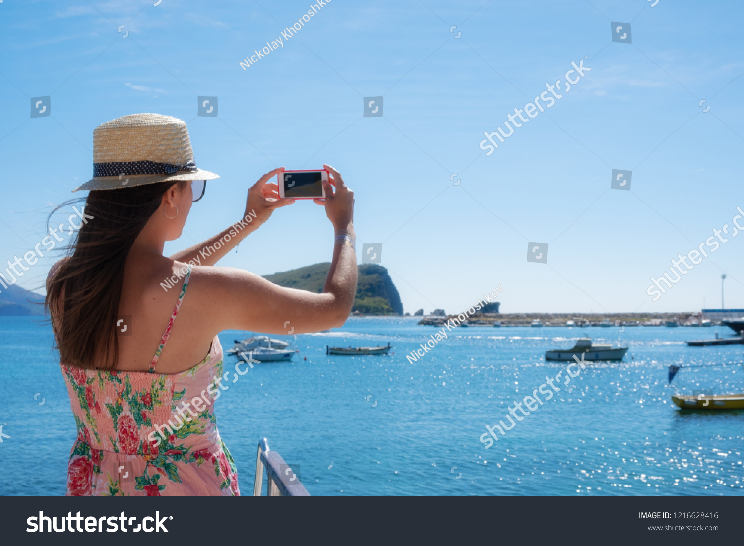 Young travelling woman in hat taking photos of summer sunny sea with smartphone camera. Brunette girl making photography on summer vacation #1216628416