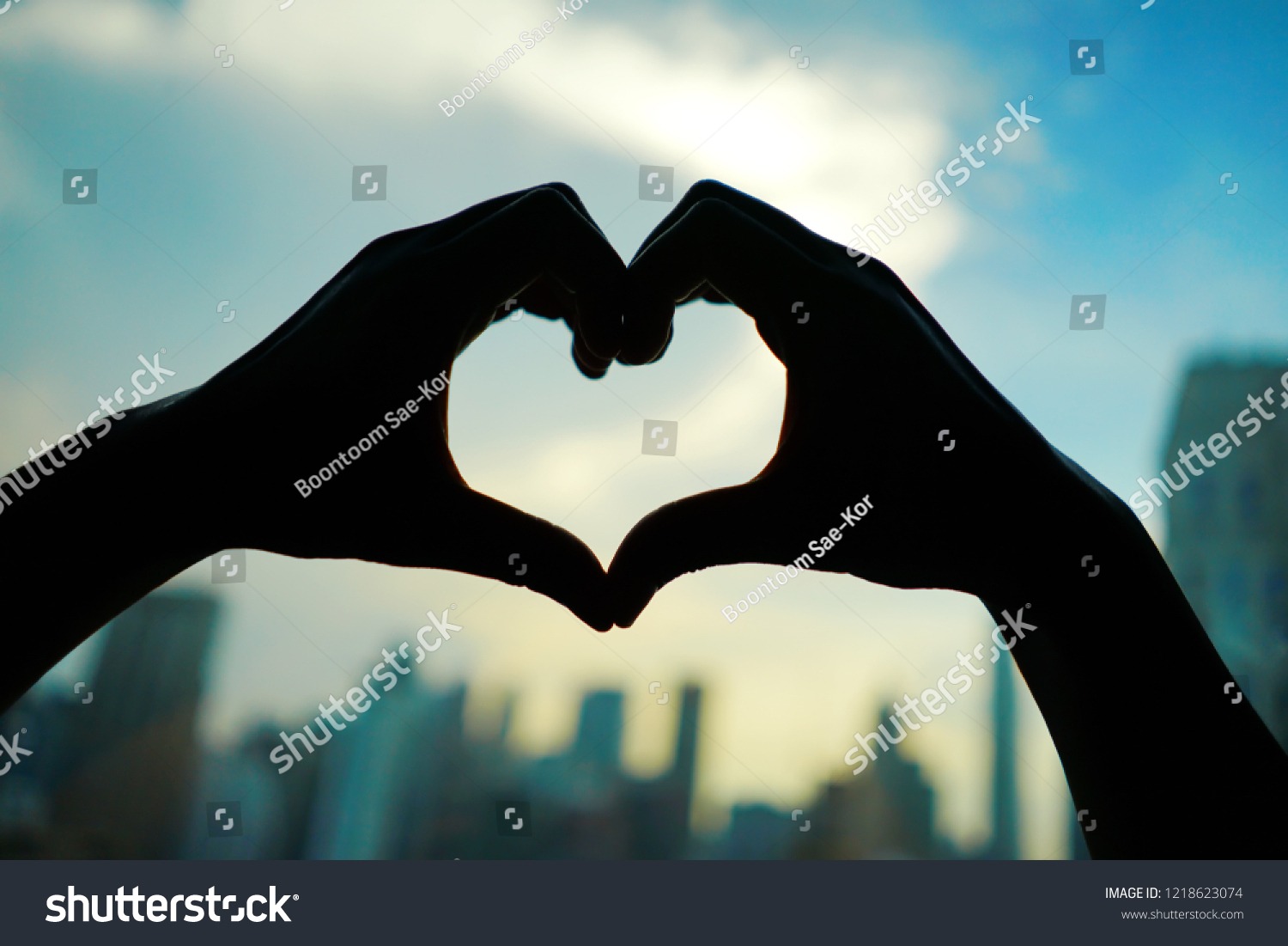 Silhouette hands making heart shaped hand gesture on blurred background of cityscape with high rise business metropolis building and skyscape with clouds at early morning or evening moment. #1218623074