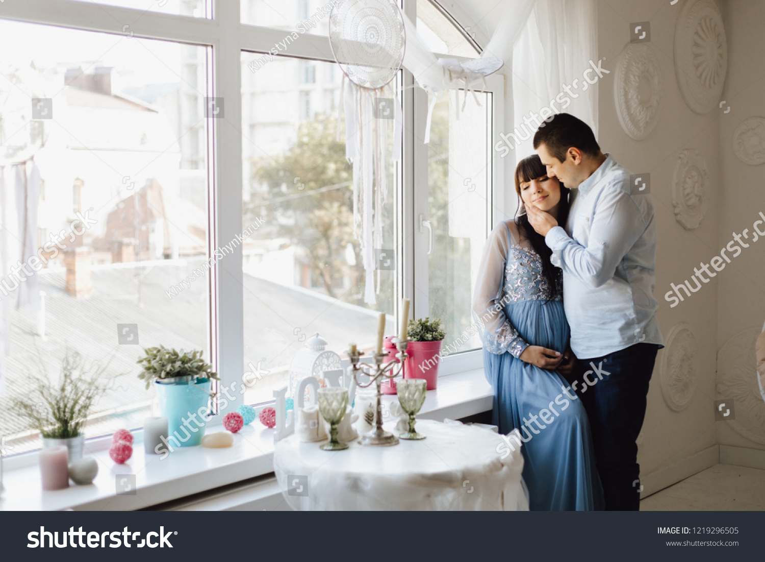 Man holds tender face of a pregnant woman while she sits on a windowsill before bright window #1219296505