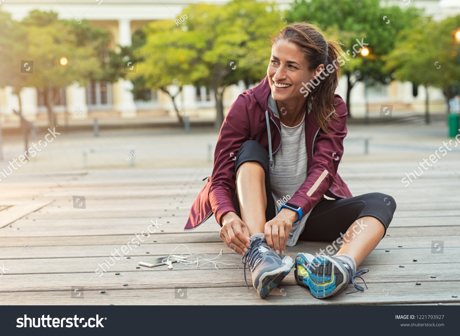 Mature fitness woman tie shoelaces on road. Cheerful runner sitting on floor on city streets with mobile and earphones wearing sport shoes. Active latin woman tying shoe lace before running. #1221793927