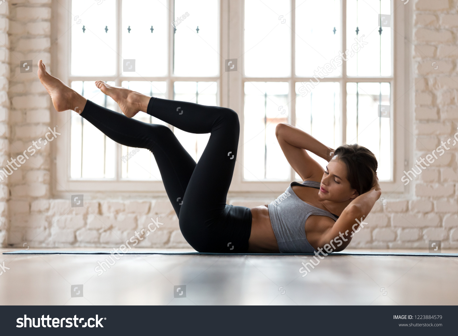 Young sporty woman practicing, doing crisscross exercise, bicycle crunches pose, working out, wearing sportswear, black pants and top, indoor full length, white sport studio #1223884579