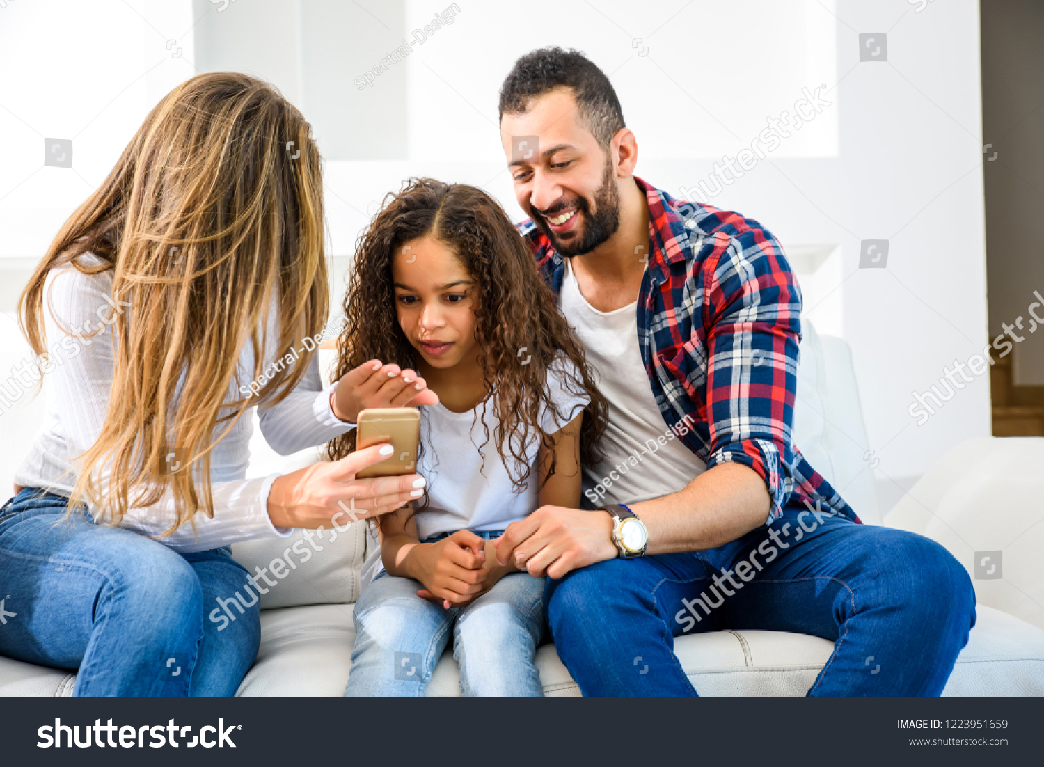 Young parents sitting on the couch with their daughter and all of them looking at a smartphone
 #1223951659