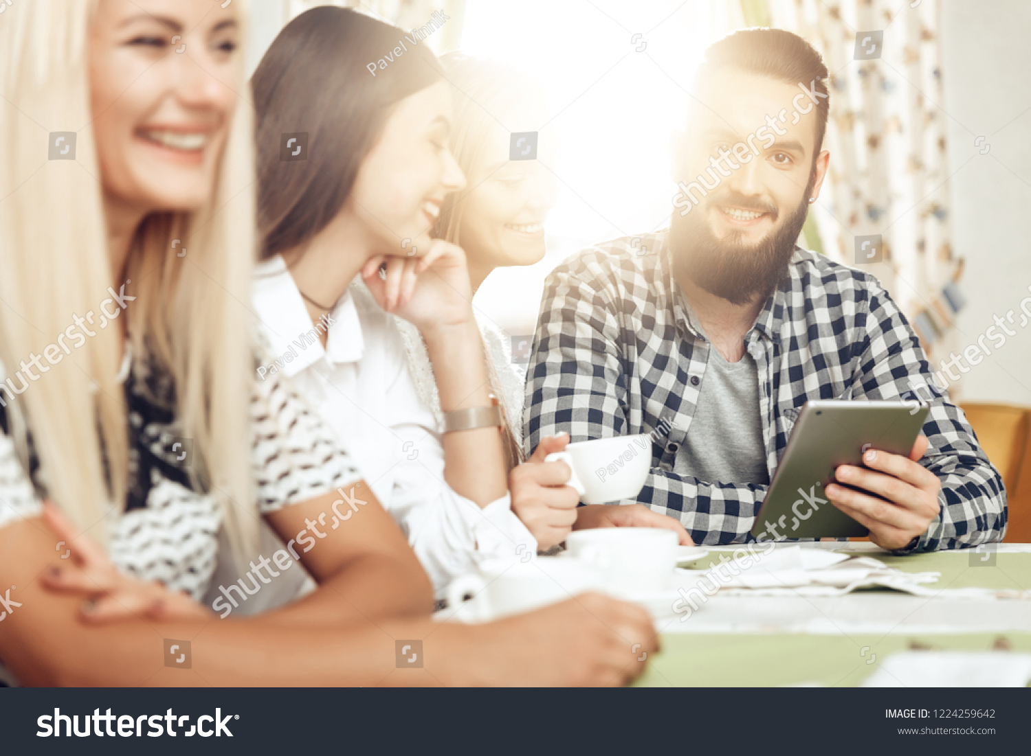 Happy friends are resting in cafe drinking coffee. A group of smiling men and women are sitting at a table in a cafe with a tablet. The guy looks at the tablet funny information that all the fun #1224259642