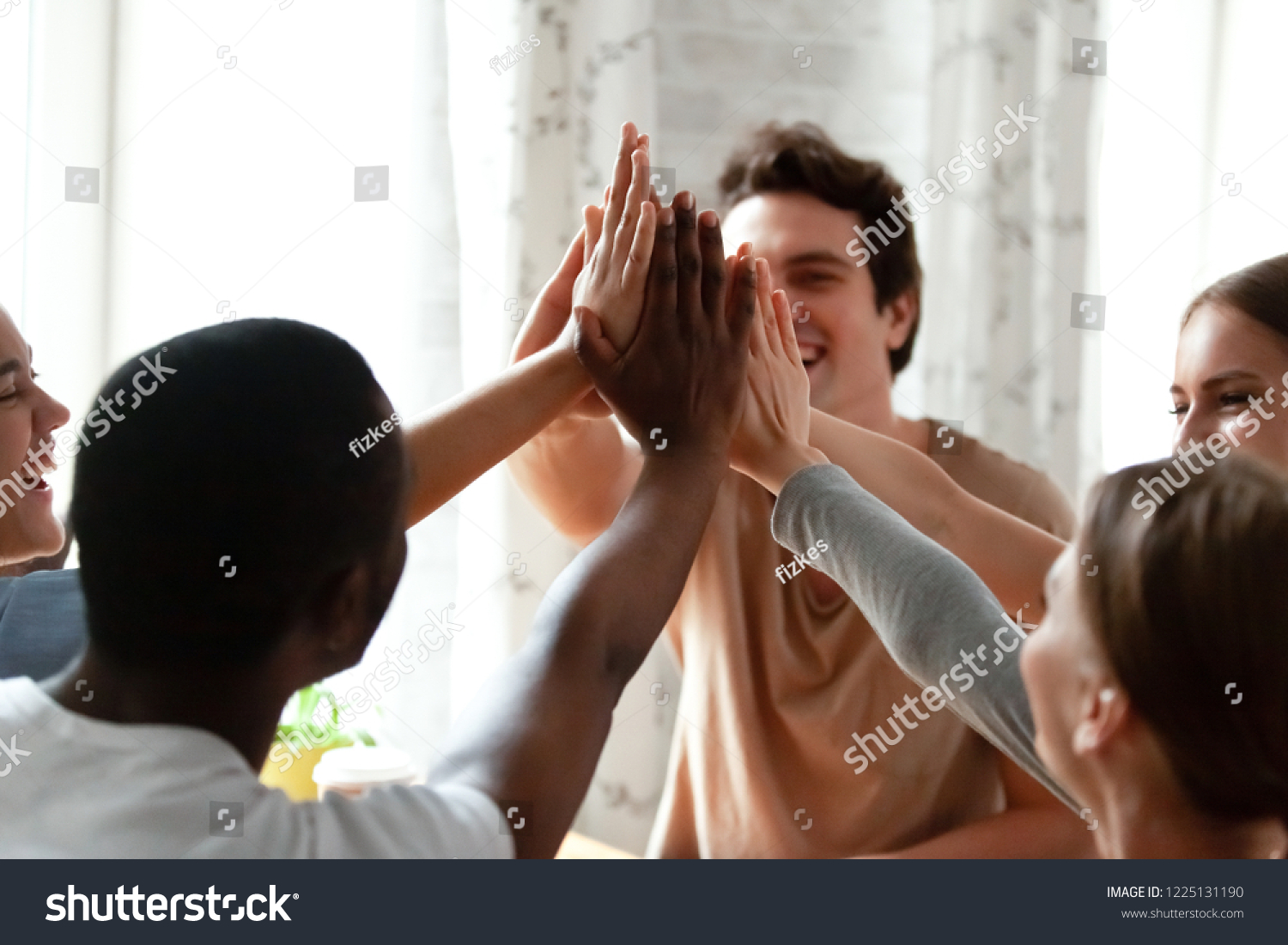 Diverse multiracial cheerful students giving high five greeting each other. Multi-ethnic millennial group of young people slapping palms sitting indoors. Gesture of celebration, friendship and unity #1225131190