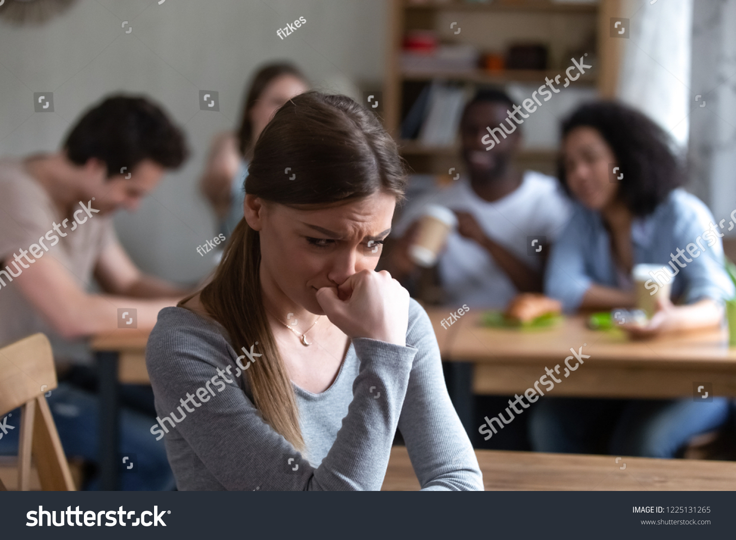 Diverse multi-ethnic friends sitting together in cafe talking having fun, focus on frustrated shy girl sitting separately by others teenagers feels unhappy because peers not accept her she is outcast #1225131265