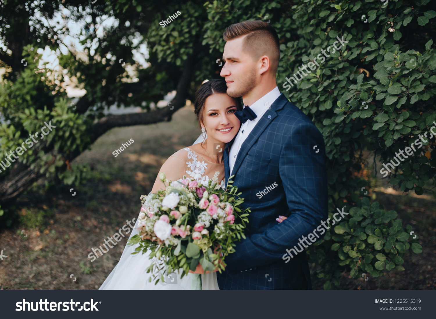 Beautiful newlyweds cuddle in a green garden with trees. Young bride and cute bride with a bouquet are standing in the park. Wedding photography. Stylish portrait. #1225515319