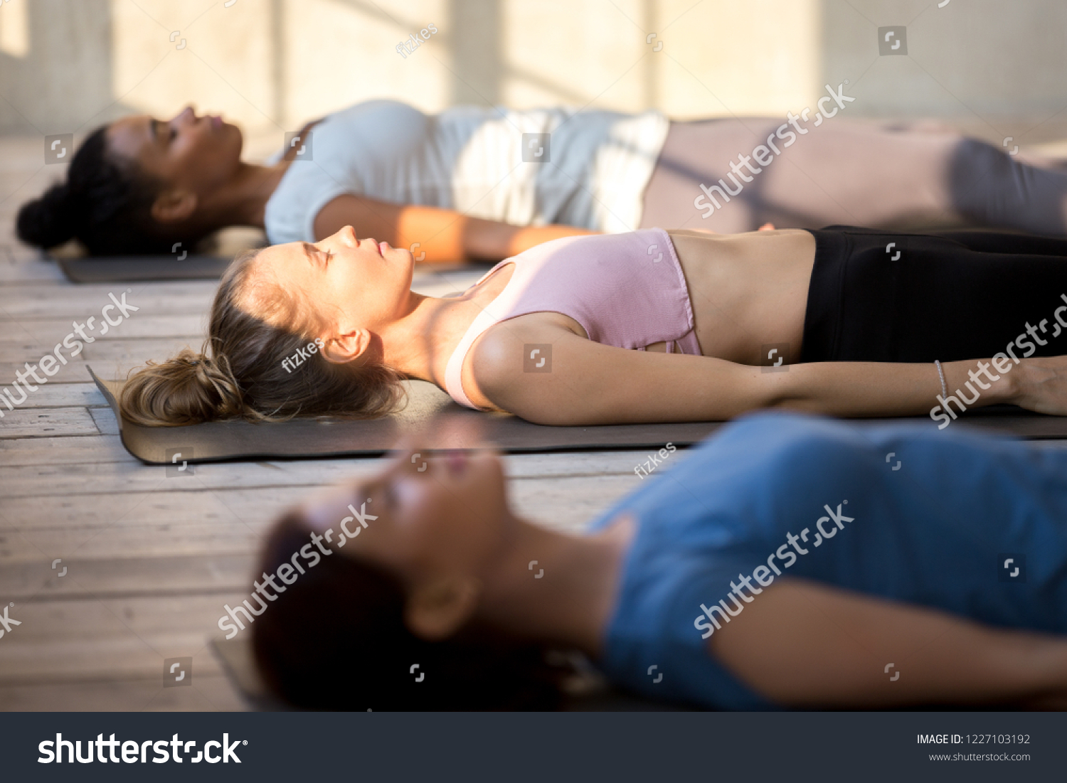 Group of young people practicing yoga lesson doing Dead Body, Savasana exercise, Corpse pose, working out, indoor close up, mixed race female students training at yoga studio. Well-being concept #1227103192