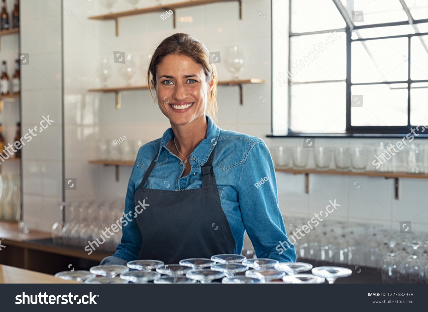 Smiling proud bartender woman standing behind counter and looking at camera. Portrait of beautiful weitress wearing denim shirt with apron. Successful small business owner at pub or coffee shop. #1227682978