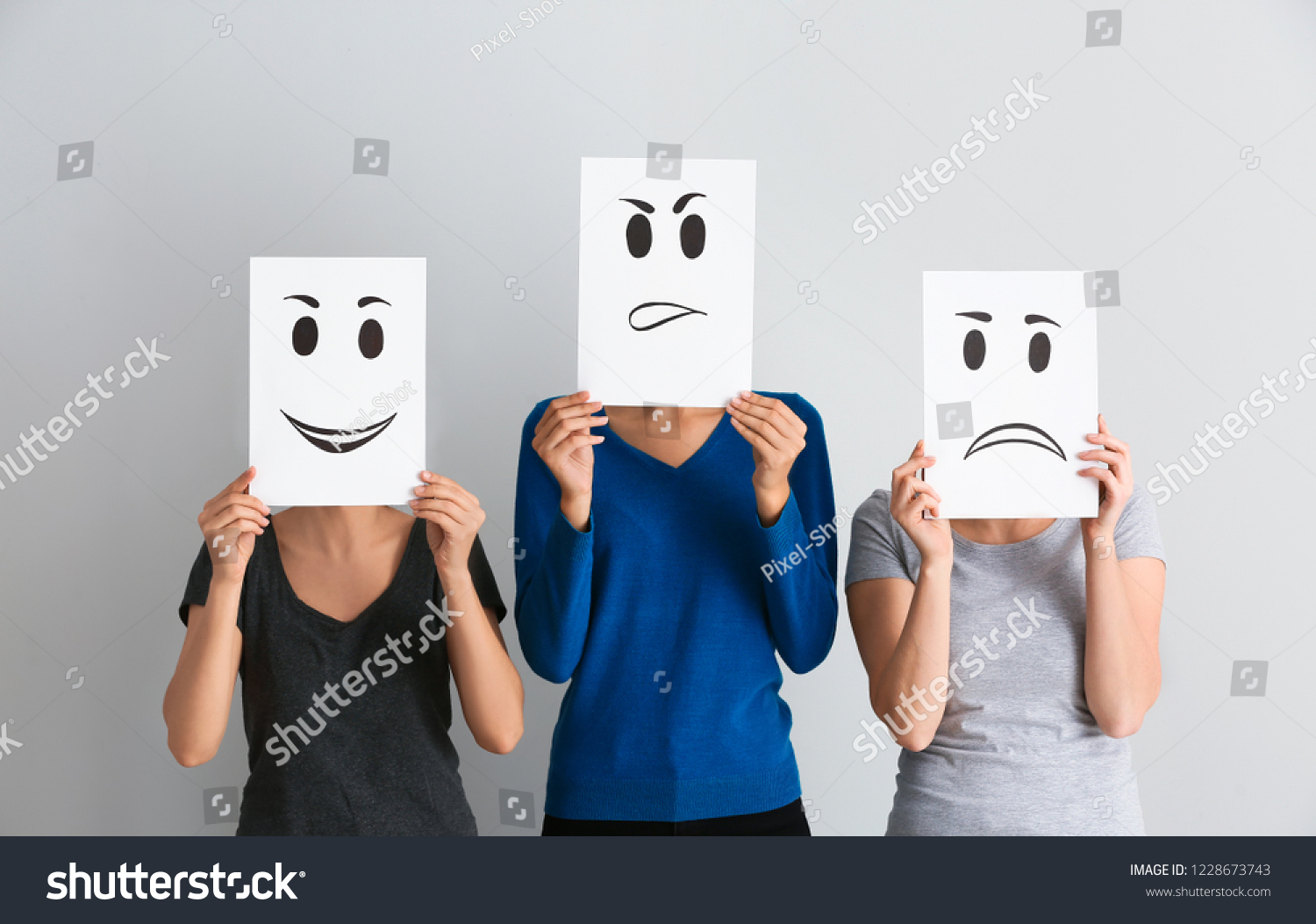 Young women hiding faces behind sheets of paper with drawn emoticons on light background #1228673743