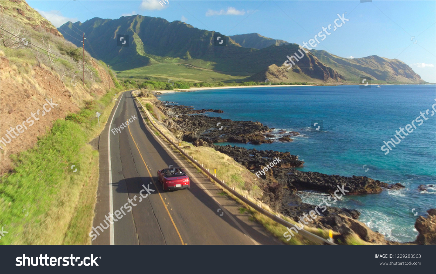 AERIAL: Red convertible car driving along the picturesque coastal road above dramatic rocky shore towards beautiful volcanic mountains. Happy young couple on summer vacation traveling at the seaside #1229288563