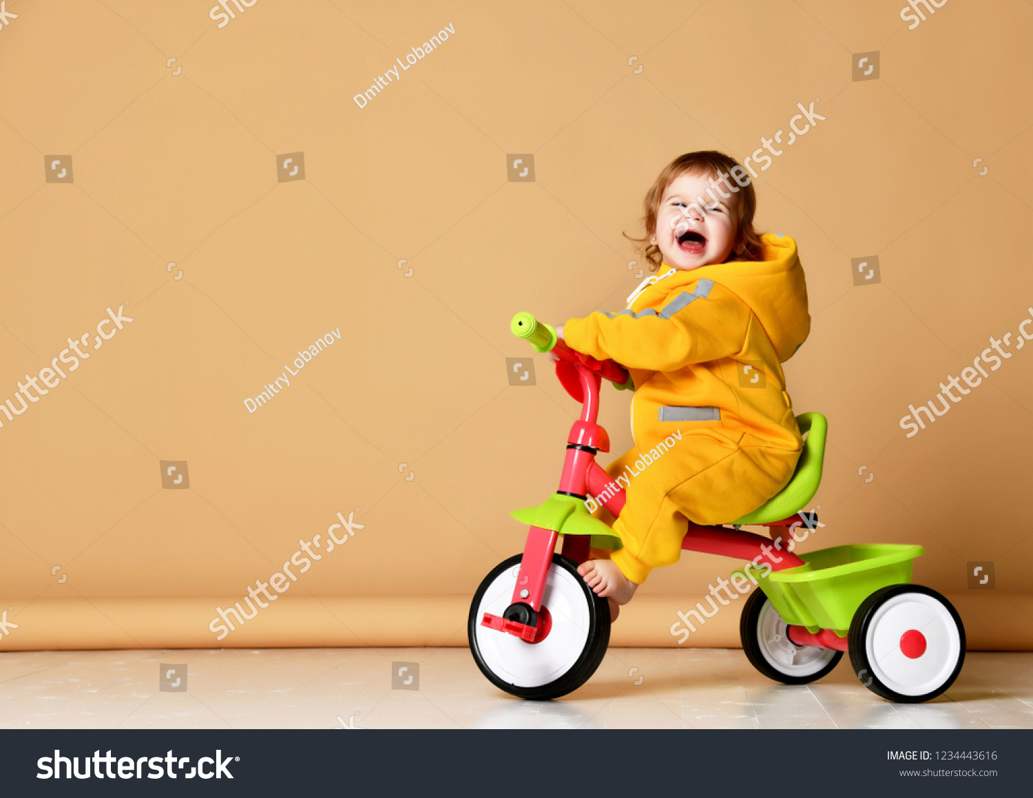 Baby girl kid riding her first bicycle tricycle in warm yellow overalls looking up on screaming grey background #1234443616