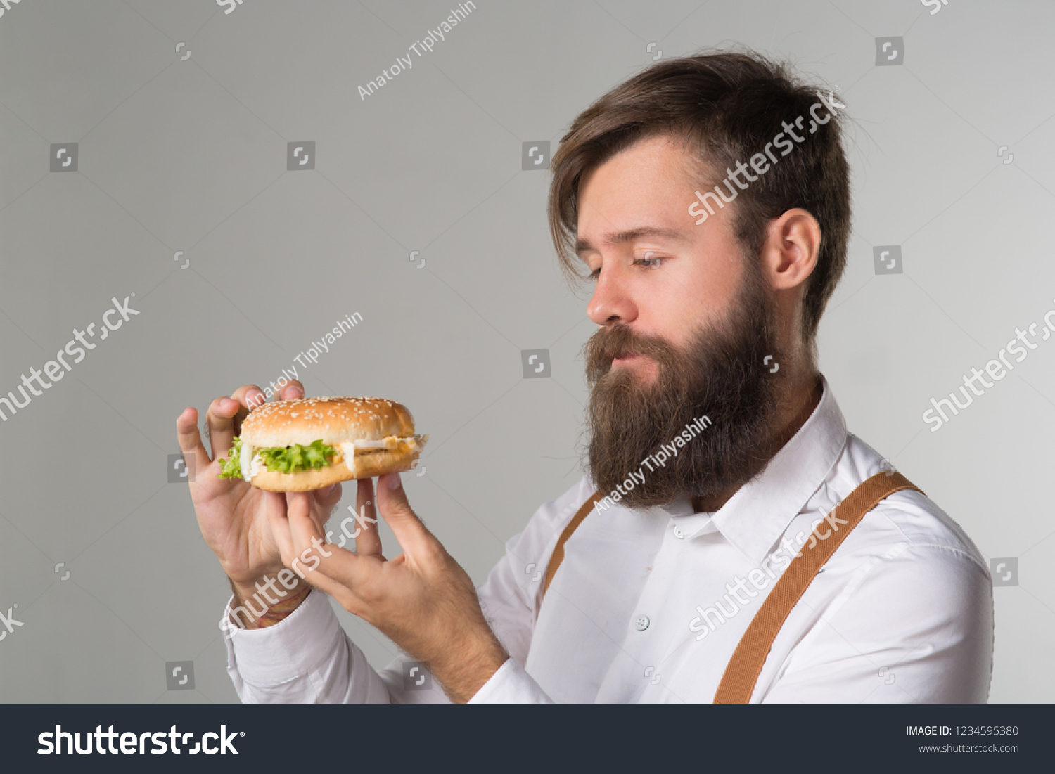 Man with beard in white shirt and suspenders eating junk food from a fast food hamburger or cheeseburger on gray background #1234595380