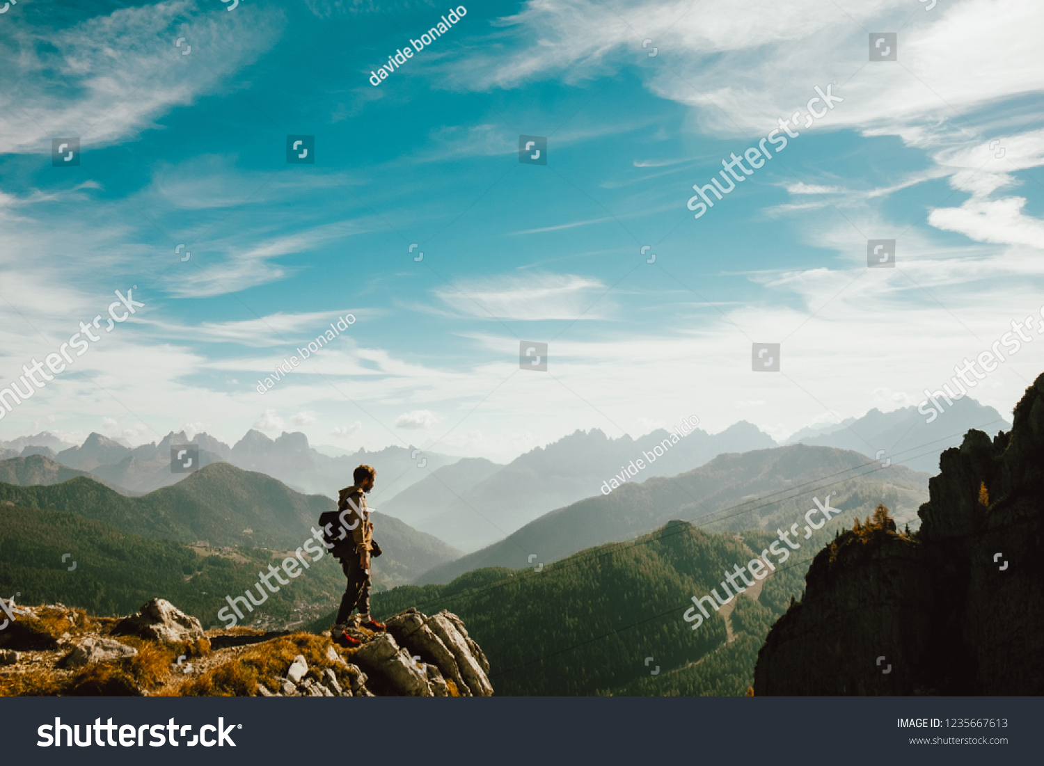 Belluno, Italy - 20 october 2018: - a young man standing over top of mountain canyon watches dramatic big valley extend in front of him making a step forward #1235667613