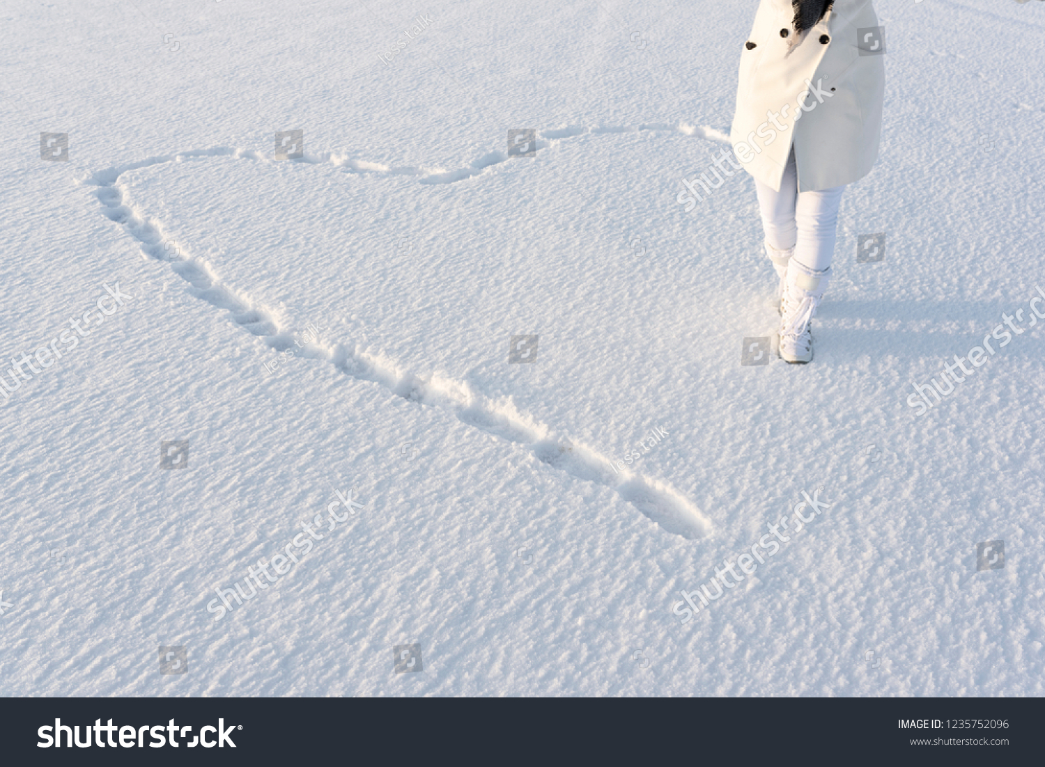 Snow heart made by footsteps. Anonymous woman dressed in white winter coat, walks on deep snow in snow boots. #1235752096