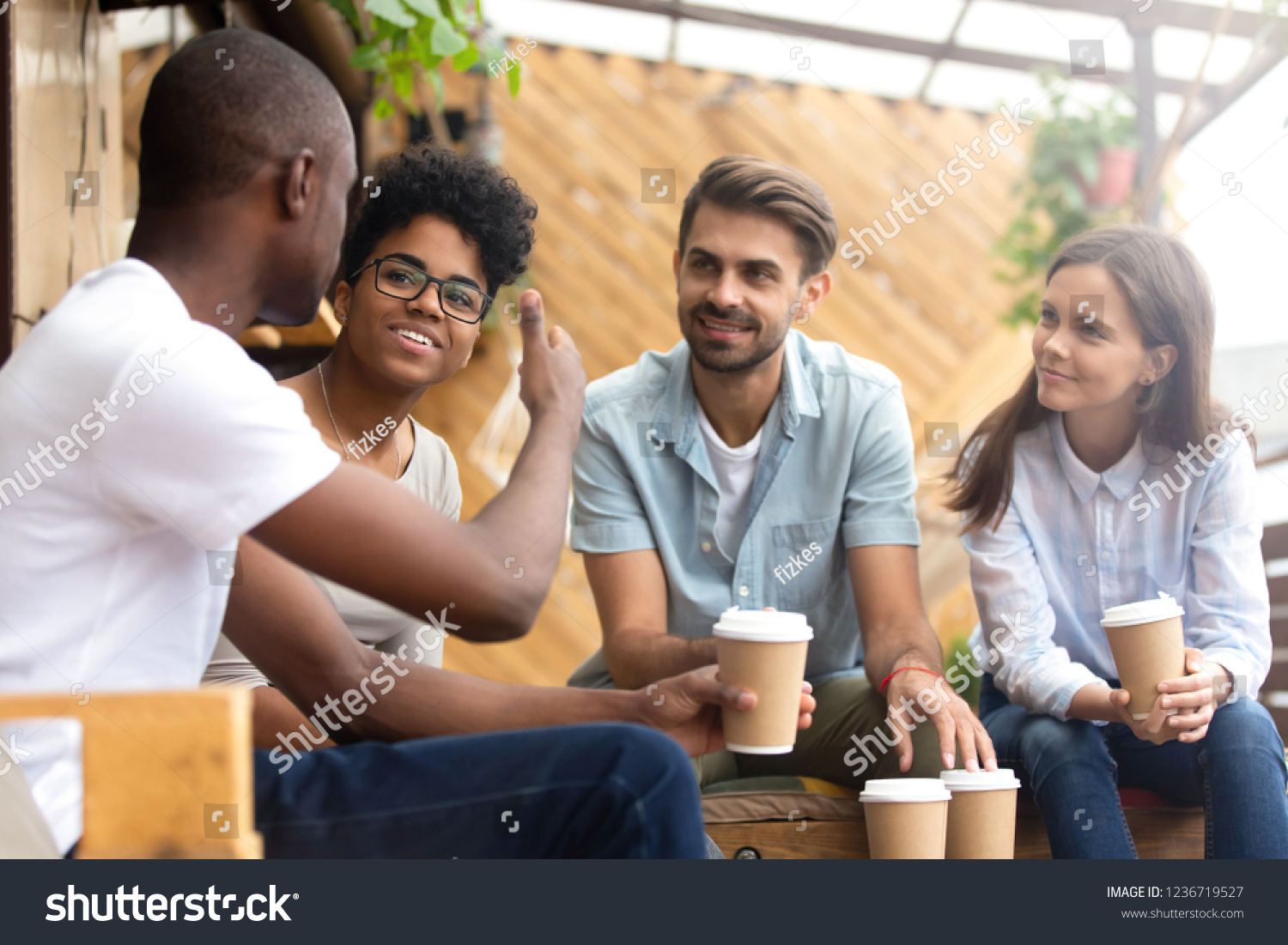 African American man showing thumb up, telling story to interested attentive friends, sitting, drinking coffee in cafe together, smiling multiethnic friends discussing, talking, chatting #1236719527