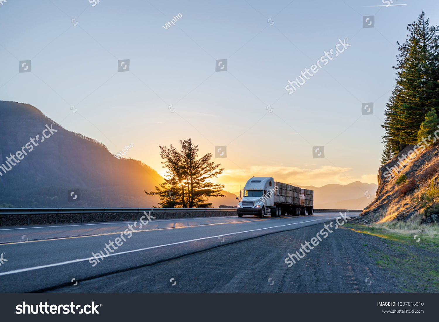 Big rig American white long haul powerful semi truck transporting boxes with fruits on flat bed semi trailer on straight evening road with scenic sunset in Columbia River Gorge #1237818910