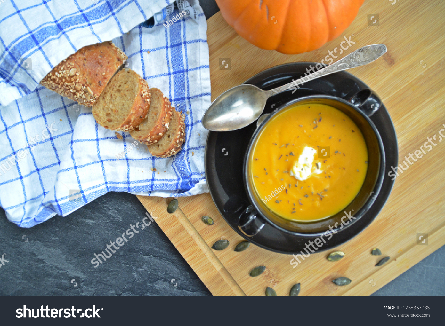 Pumpkin cream soup in a small bowl and on a wooden board on a kitchen surface with a whole pumpkin and sliced ​​bread beside it - autumnal feasting #1238357038