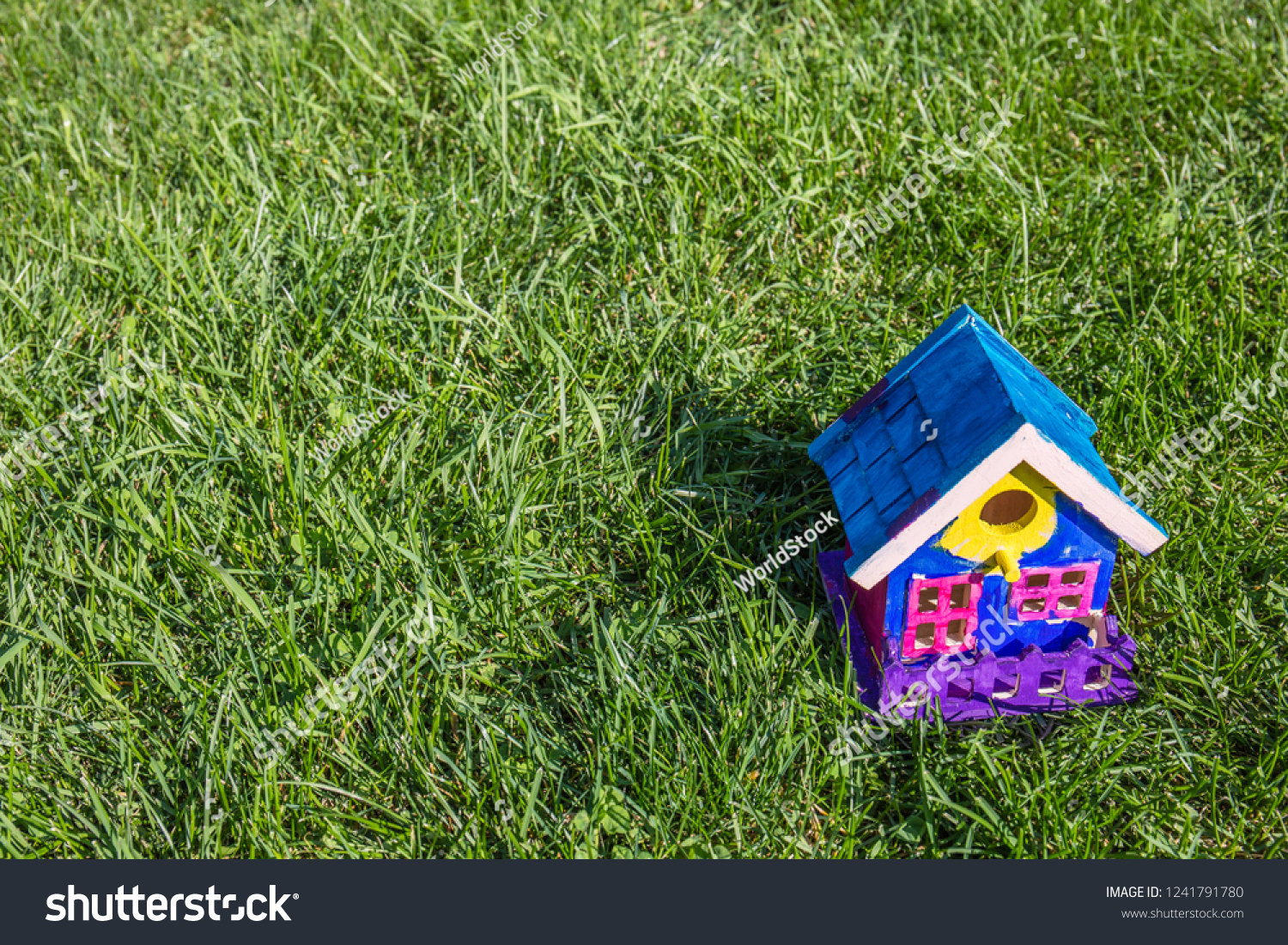 A handmade, colorful, brightly painted bird house resembling a family home in the bottom corner of a green lawn in bright sunshine. #1241791780