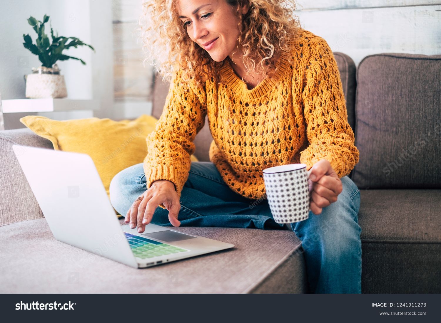 Nice beautiful lady with blonde curly hair work at the notebook sit down on the sofa at home - check on oline shops for cyber monday sales - technology woman concept for alternative office freelance #1241911273
