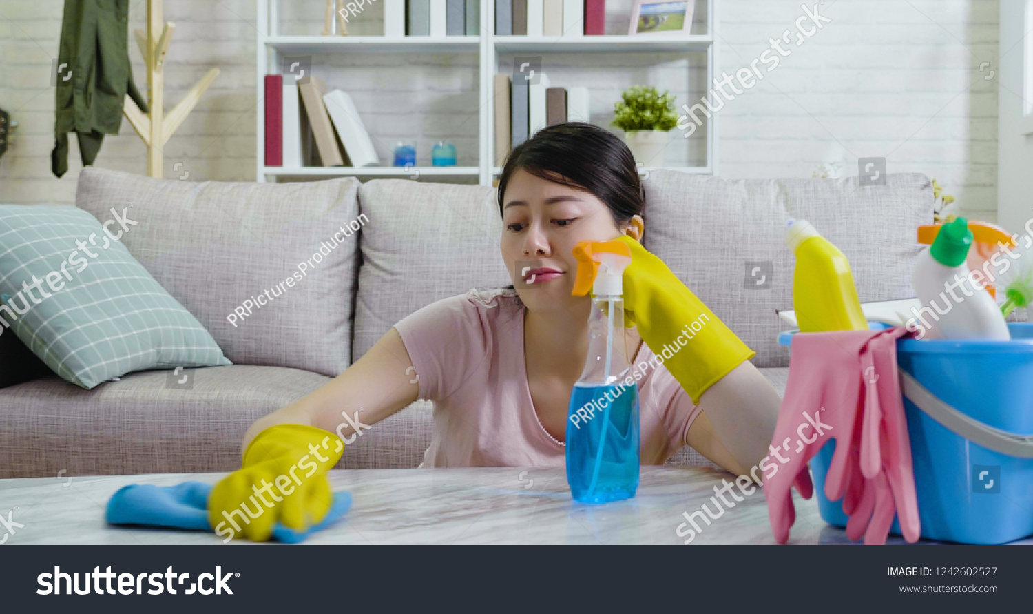 asian housewife unhappy doing home chores with hands putting under the chin while wiping the table sitting on the floor in a cozy room indoors. lady in protective gloves with unsatisfied face. #1242602527