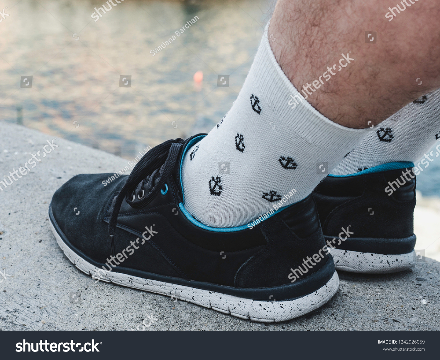 Men's legs in sports shoes and white socks with a bright, sea pattern in the shape of anchors on the background of a rocky shore on a summer, sunny day. Concept of fashion, sport and recreation #1242926059