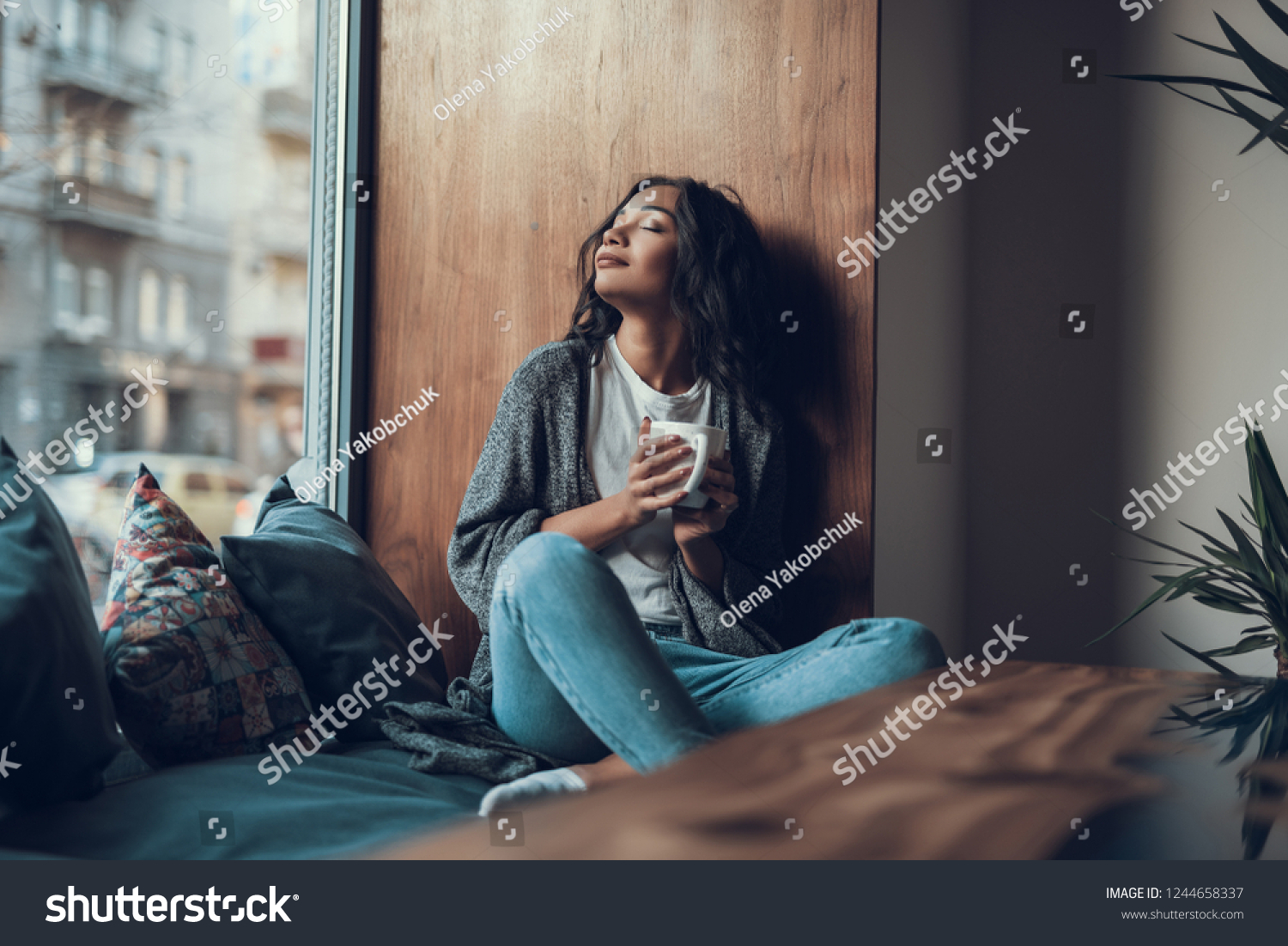 Peaceful young lady sitting on the window sill and smiling while drinking tea with her eyes closed #1244658337