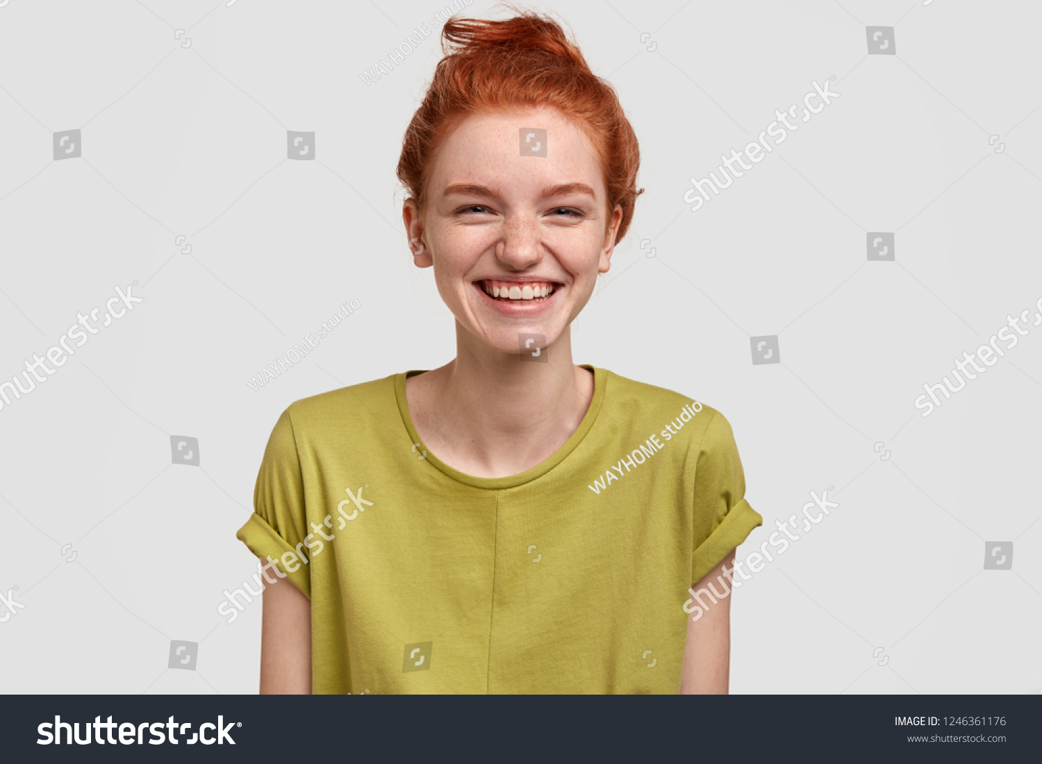 Lovely red haired girl with positive expression, laughs as watches funny TV show, enjoys weekend, dressed in green t shirt, has freckled skin, isolated over white background, amused by comic idea #1246361176