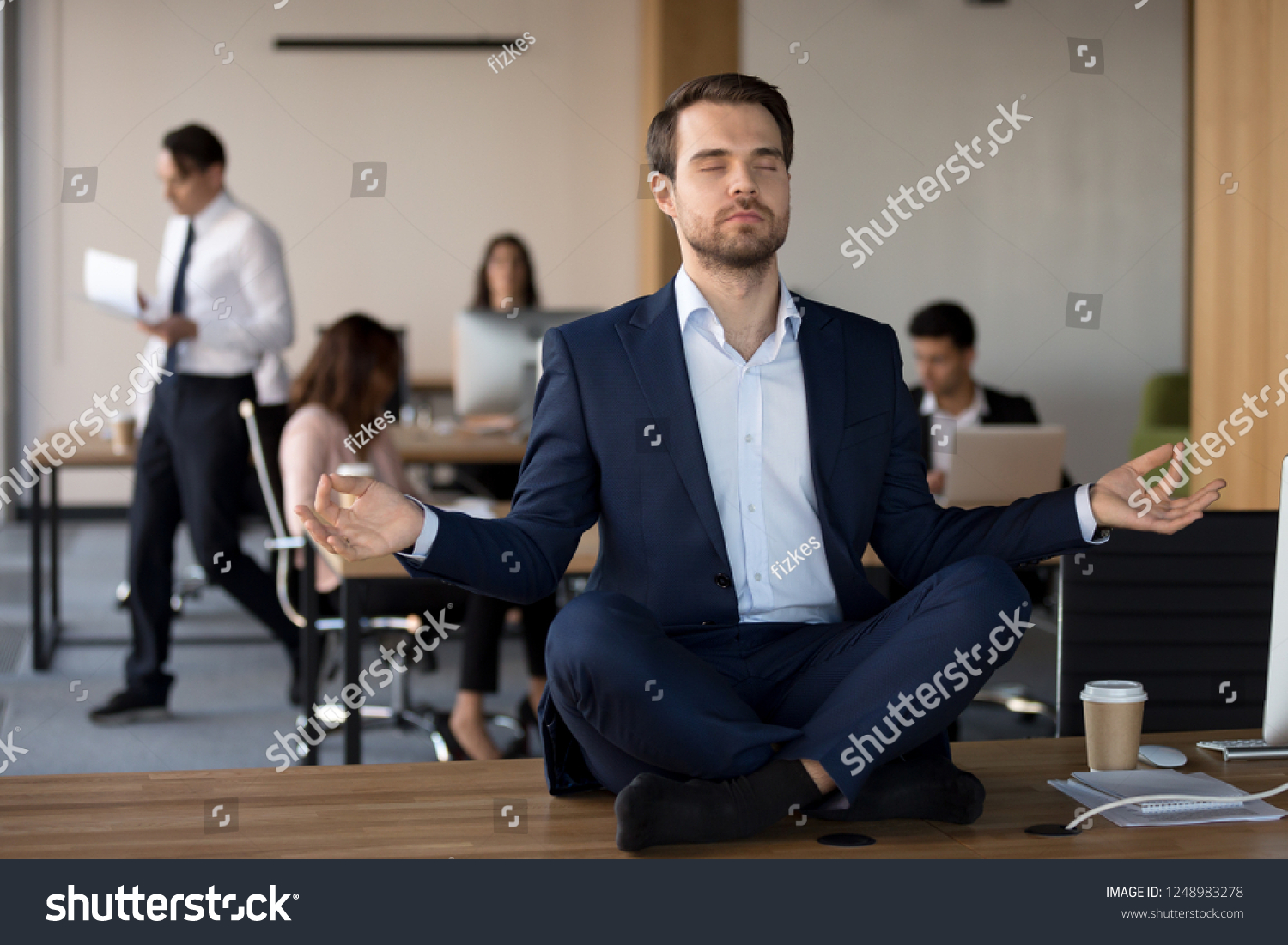 Business People working in coworking space, focus on millennial peaceful employee in formal suit sitting without shoes in lotus position on office desk practising meditation and visualization exercise #1248983278