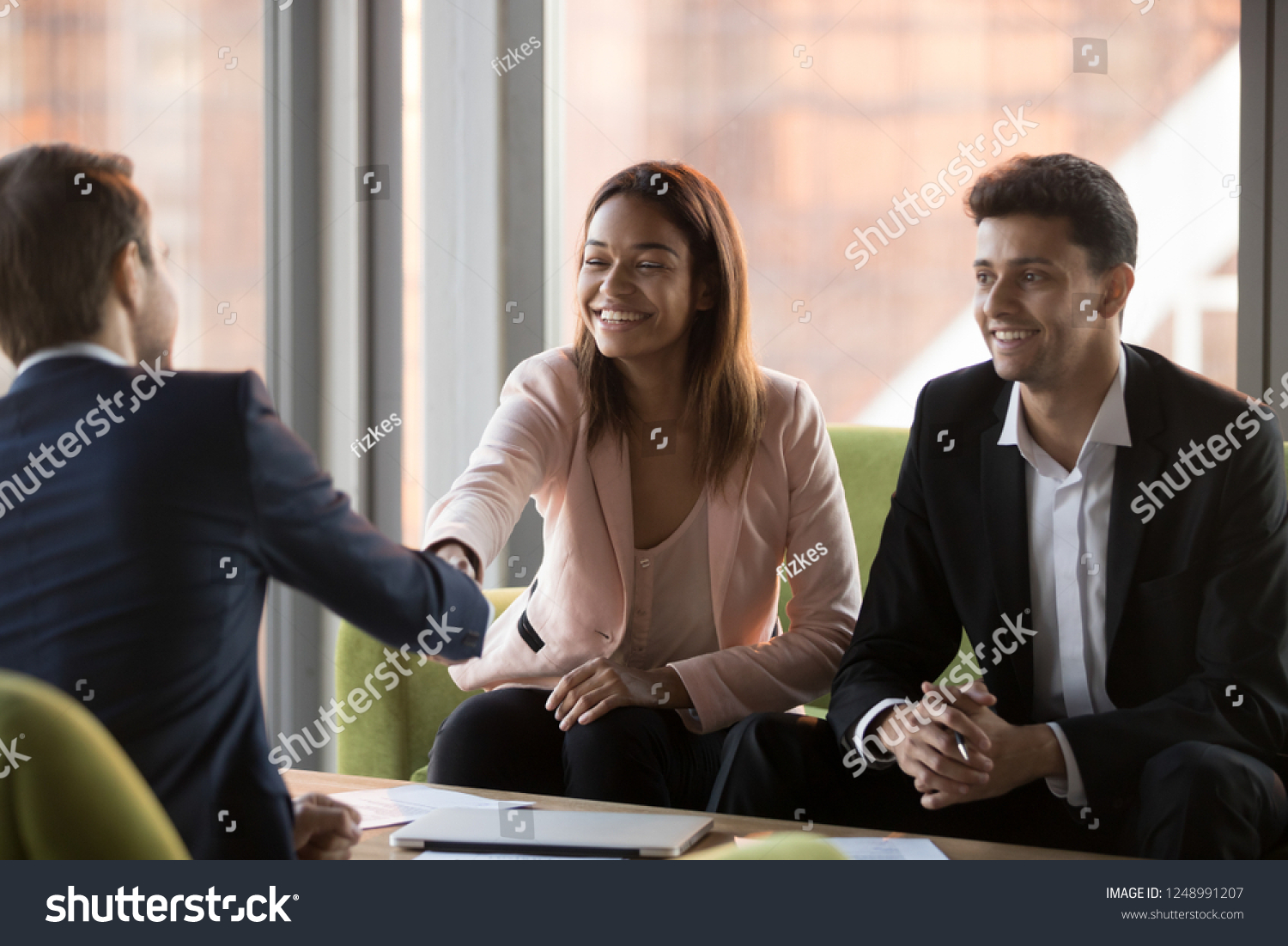 Positive millennial middle eastern ethnicity businessman and african american businesswoman greeting company client, customer rear view, sitting on couches shaking hands ready to start conversation #1248991207
