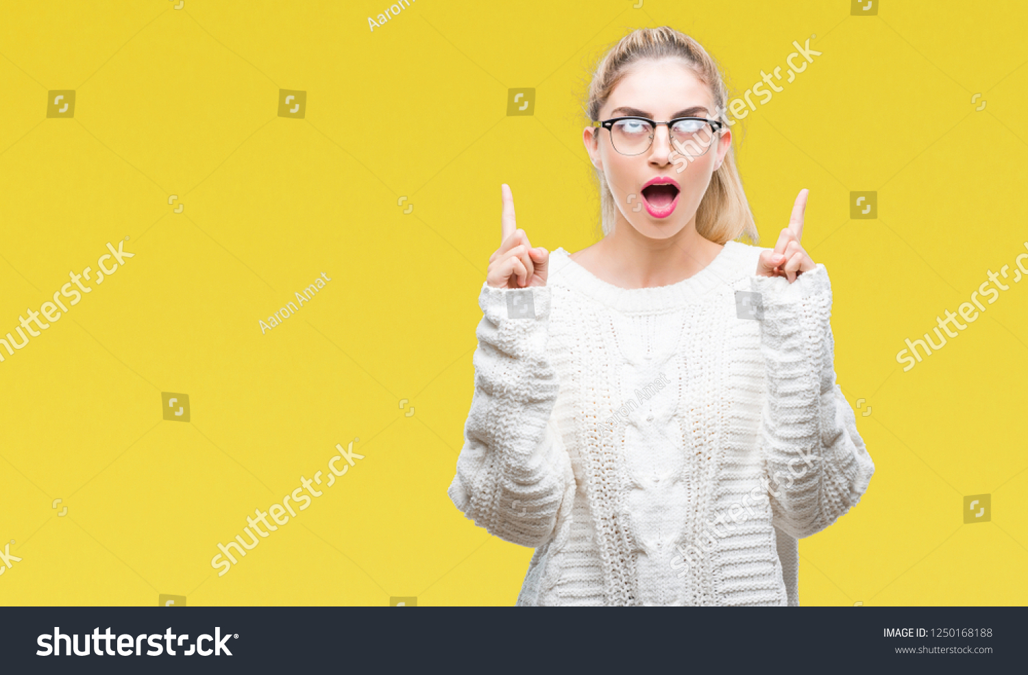 Young beautiful blonde woman wearing glasses over isolated background amazed and surprised looking up and pointing with fingers and raised arms. #1250168188