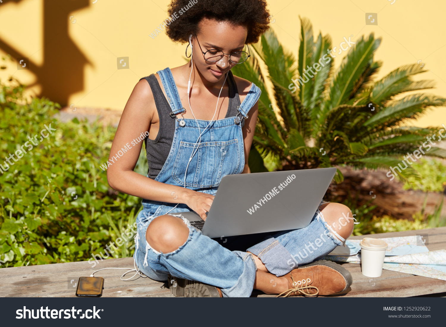 Photo of attractive woman with Afro haircut, sits crossed legs with portable laptop computer, keyboards new publication for her blog, listens music, uses mobile phone, earphones, wears stylish clothes #1252920622