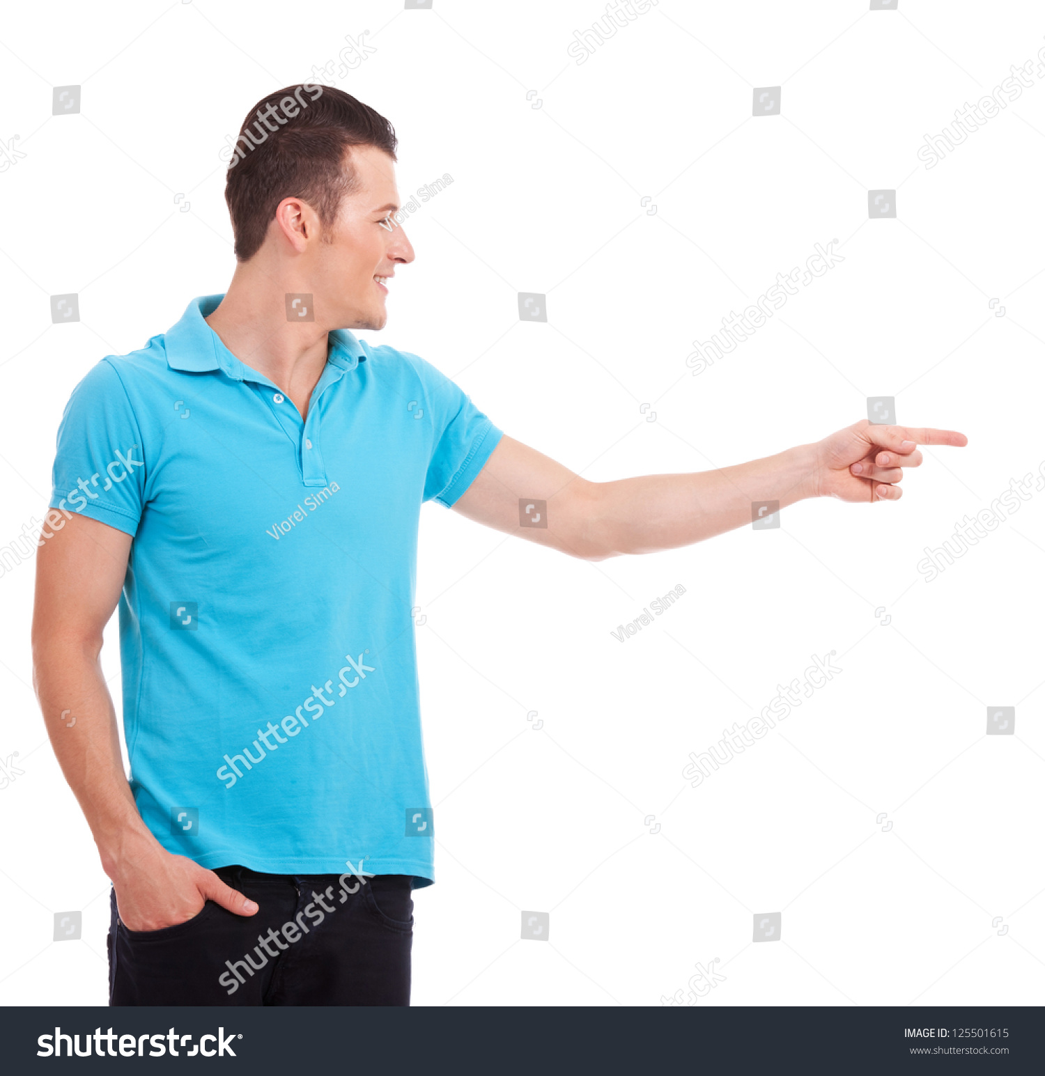 Casual young man pointing and looking to his side, with the other hand in his pocket, over white background #125501615