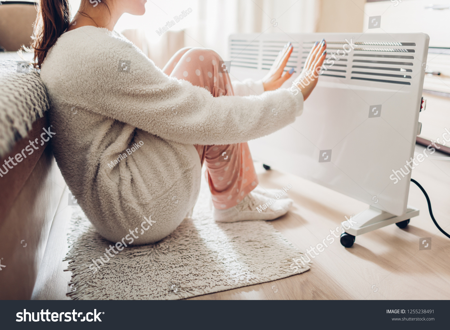 Using heater at home in winter. Woman warming her hands sitting by device and wearing warm clothes. Heating season. #1255238491