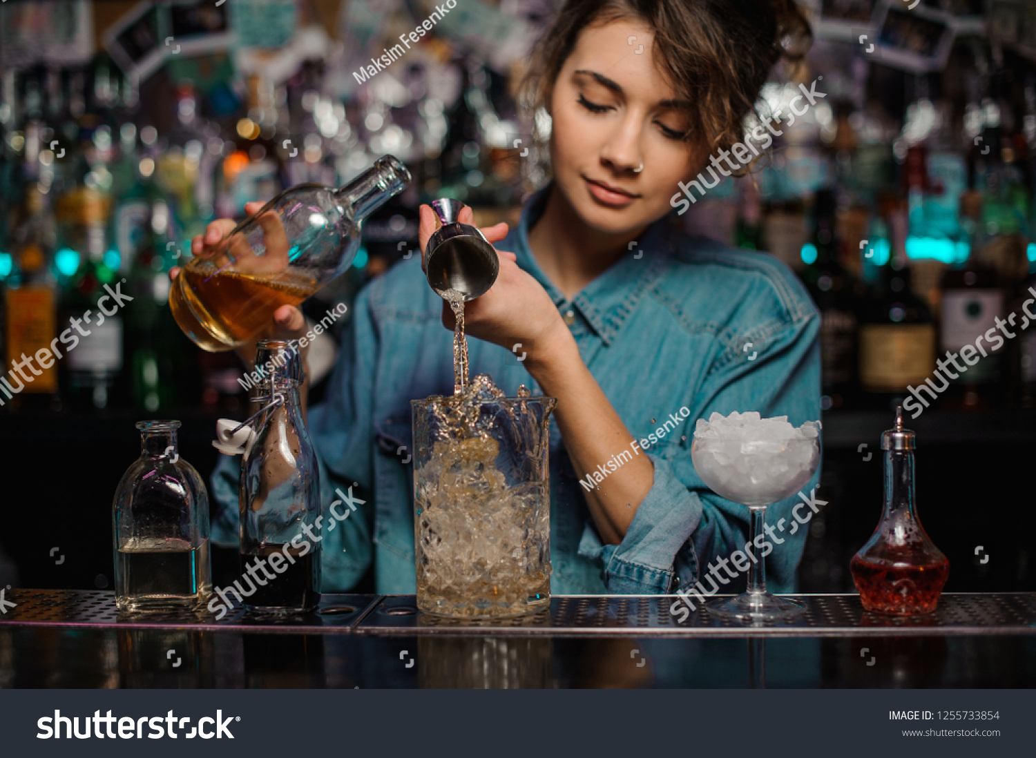 Female bartender pouring to the measuring glass cup with ice cubes an alcoholic drink from steel jigger on the bar counter on the blurred background #1255733854