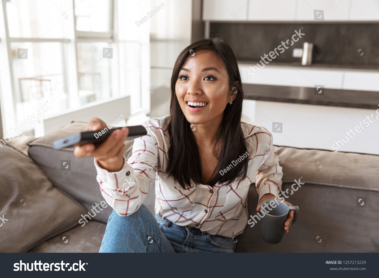 Photo of positive asian woman 20s holding remote control and watching TV while sitting at sofa in cozy apartment #1257213229