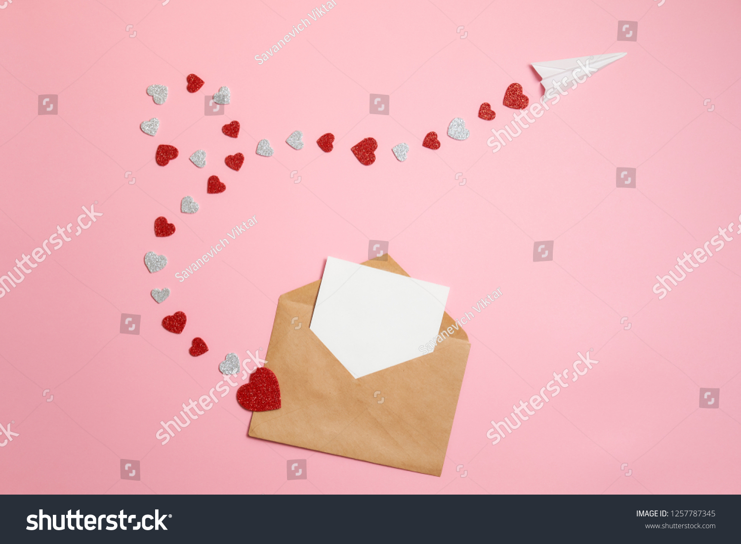 Distance love concept, sending love letter, valentines day. Kraft envelope with blank postcard and paper airplane flying on route made of heart shaped valentines cards lay on pink background desk #1257787345