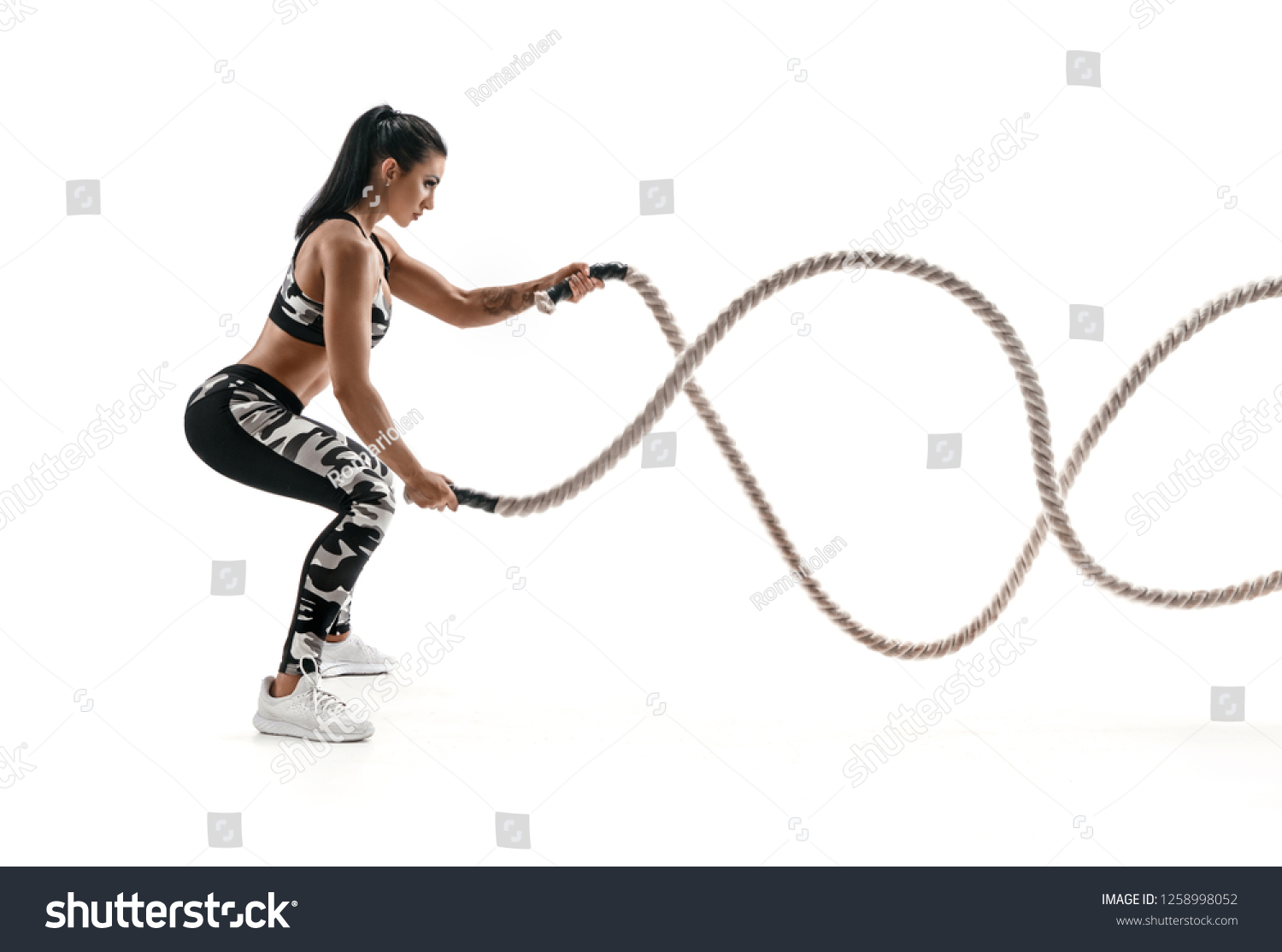Strong muscular woman working out with battle ropes. Photo of attractive woman in fashionable sportswear isolated on white background. Strength and motivation. Side view. #1258998052