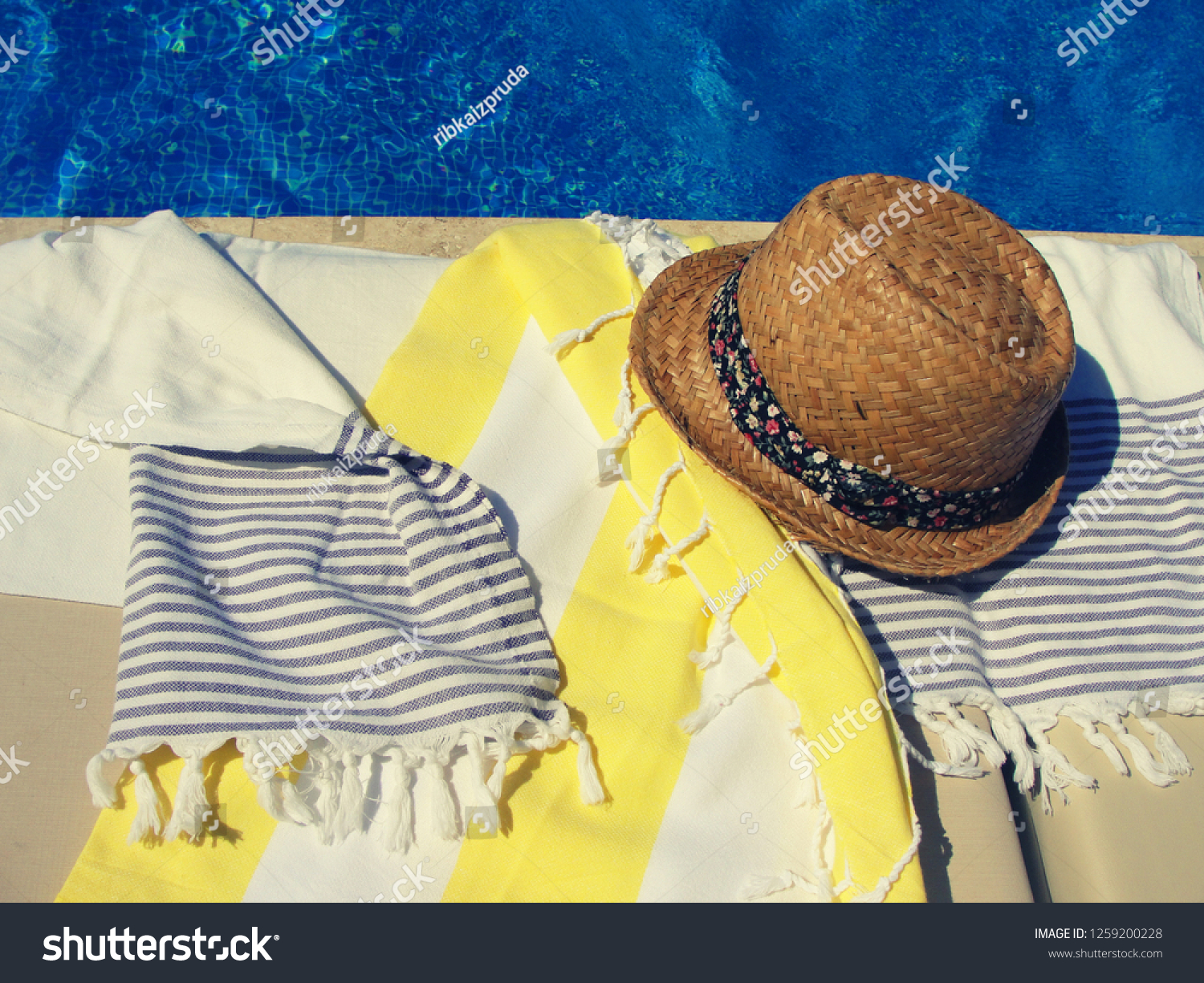 Straw summer hat with ribbon and cotton towels on a sunbed near the swimming pool. Bright sunny summer vacation day, filtered image, contrast filter effect #1259200228