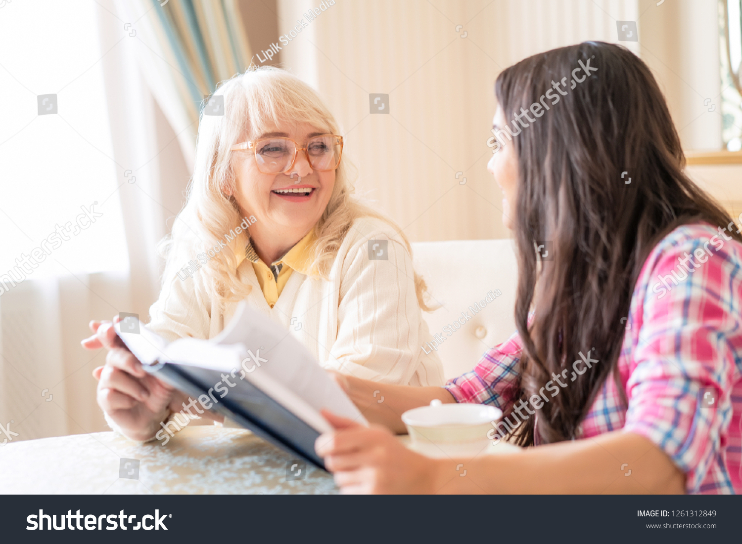 Delighted Elderly Mother Looks Happily On Her Younger Daughter Who Holds An Open Book And Drinks Tea. Caregiving Sitting. #1261312849