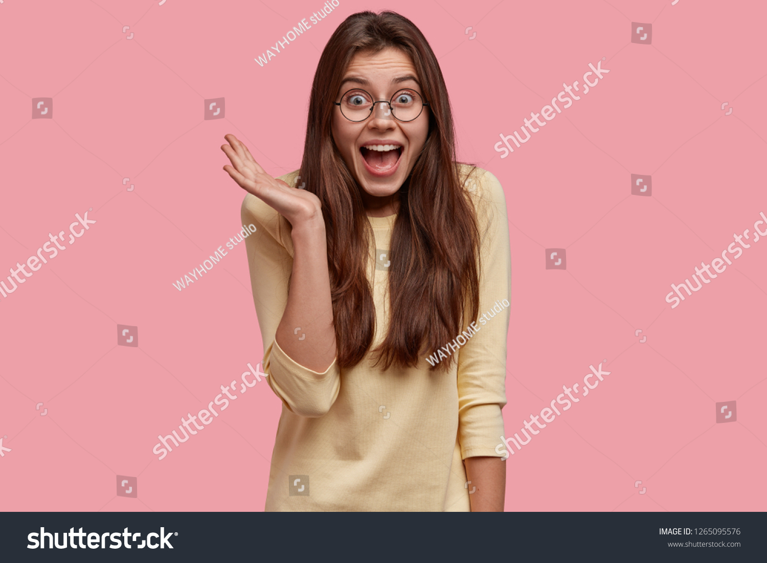 Overjoyed pretty lady keeps hand raised, exclaims with happiness, has long hair, dressed in casual outfit, isolated over pink background, expresses positive emotions. Wow, thats just amazing #1265095576