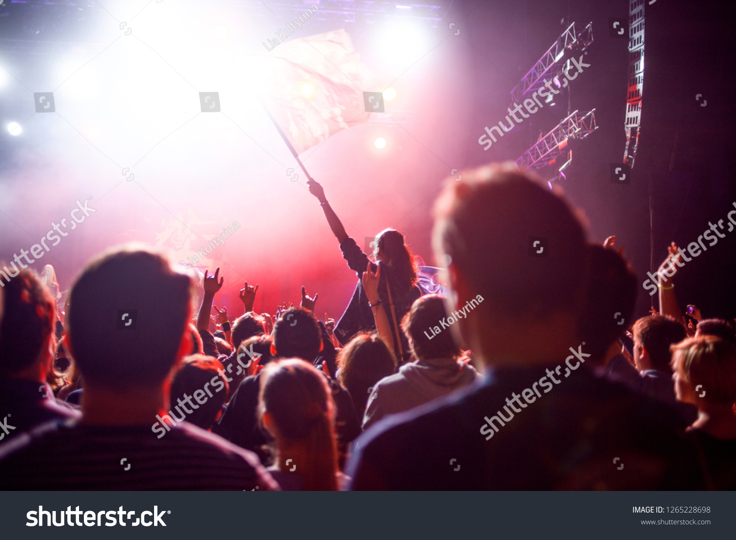 Rock concert, people in the hall on the background of the stage and spotlights. Fans waving the flag and making hands up #1265228698