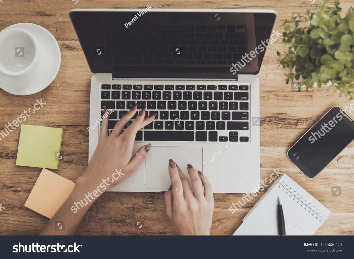 Top view of the working space of a modern business. Female hands are typed on laptop that stands next to cup and notepad on wooden desktop. Digital technologies concept. Creative space. #1265686420