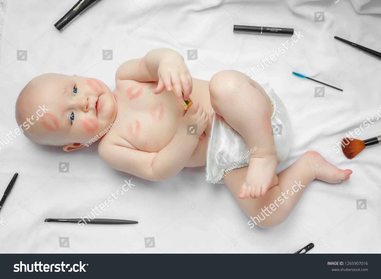 Cute happy newborn baby with red lipstick kisses on the skin with scattered makeup tools on white background. top view. #1265907016