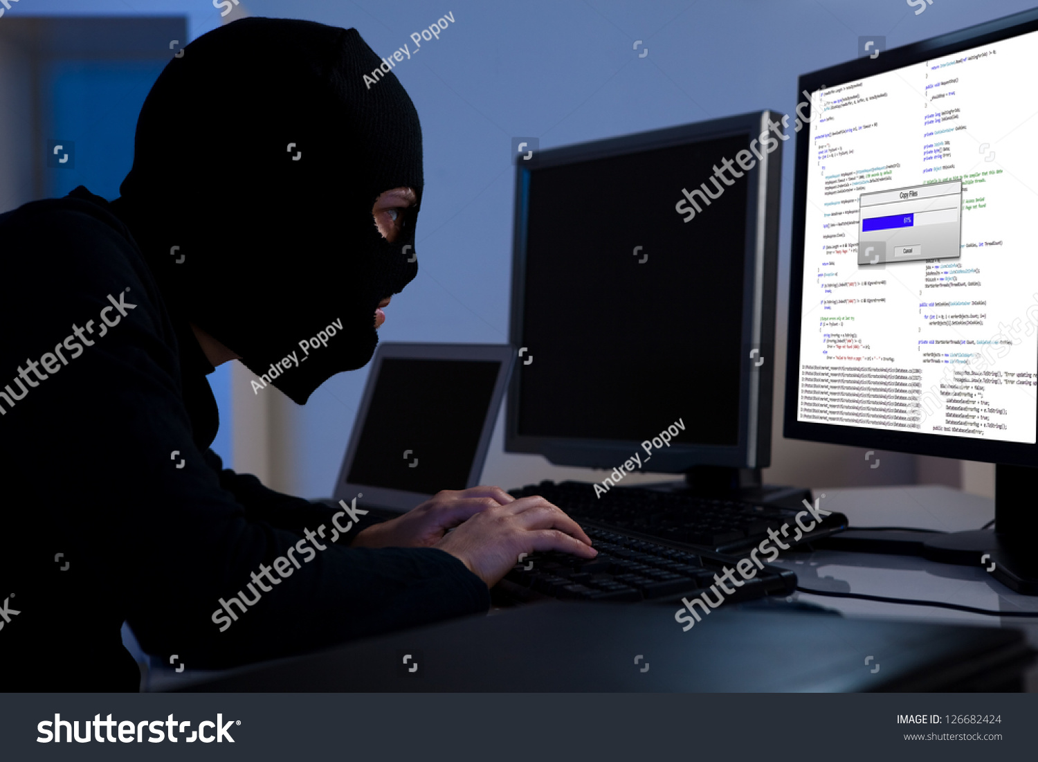 Masked hacker wearing a balaclava sitting at a desk downloading private information off a computer #126682424
